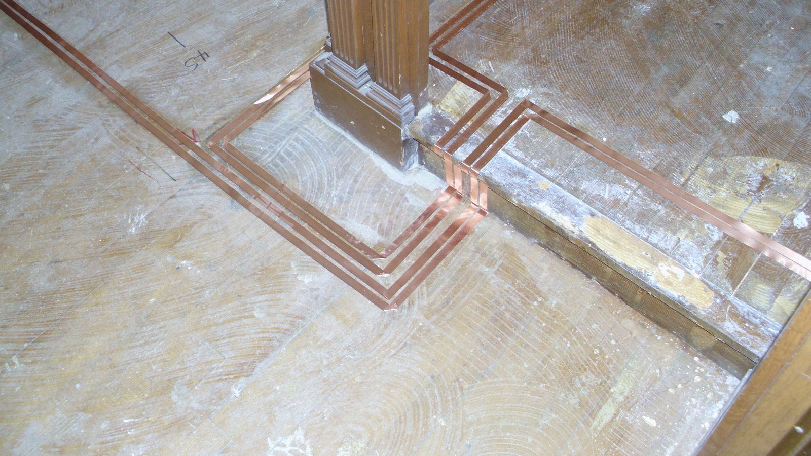 Three parallel runs of flat copper tape turning in neat right angles around a carved wooden post on a bare wood floor