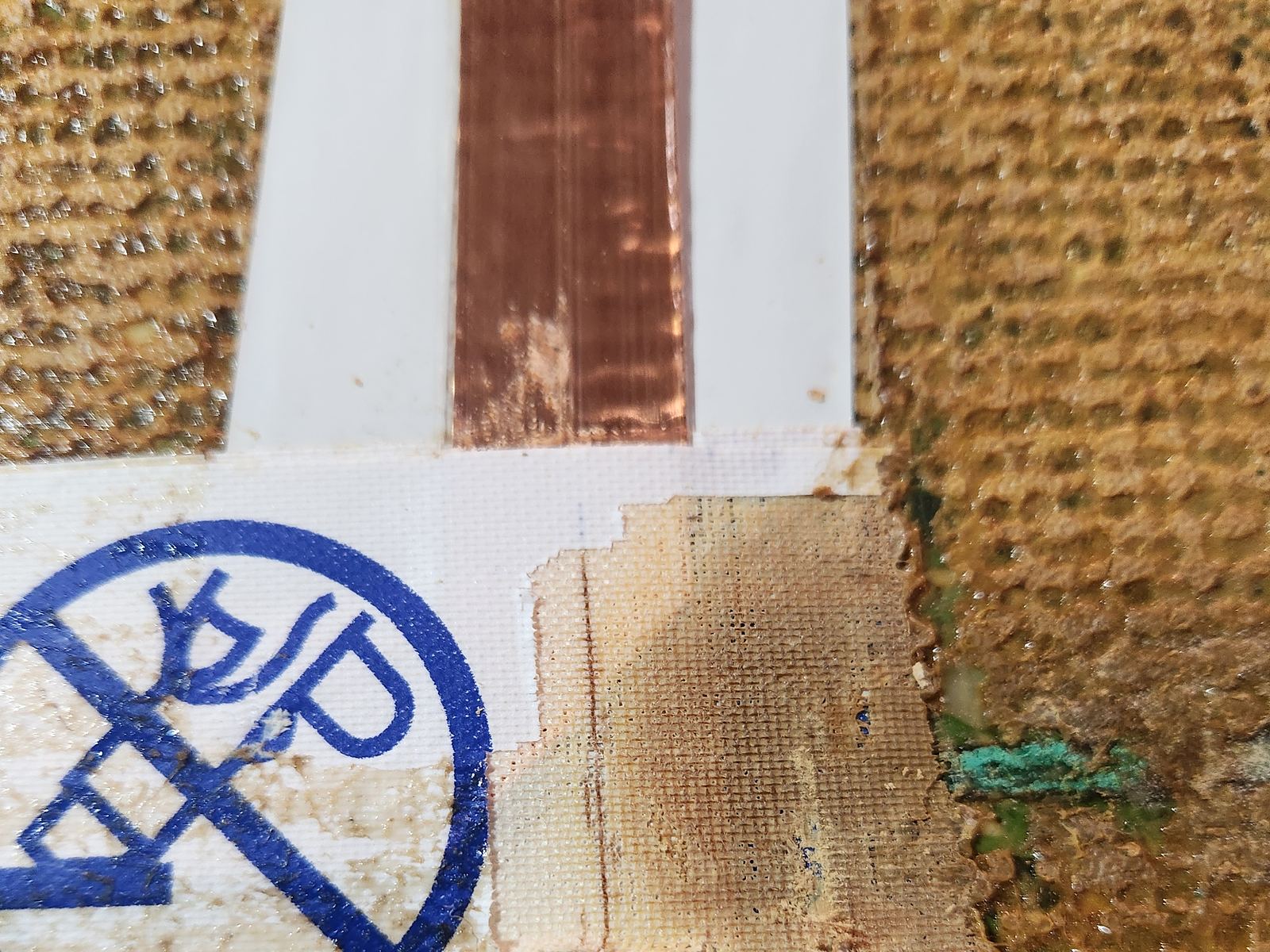 A strip of copper loop tape exposed beside rubber carpet backing, next to a printed warning label on the floor