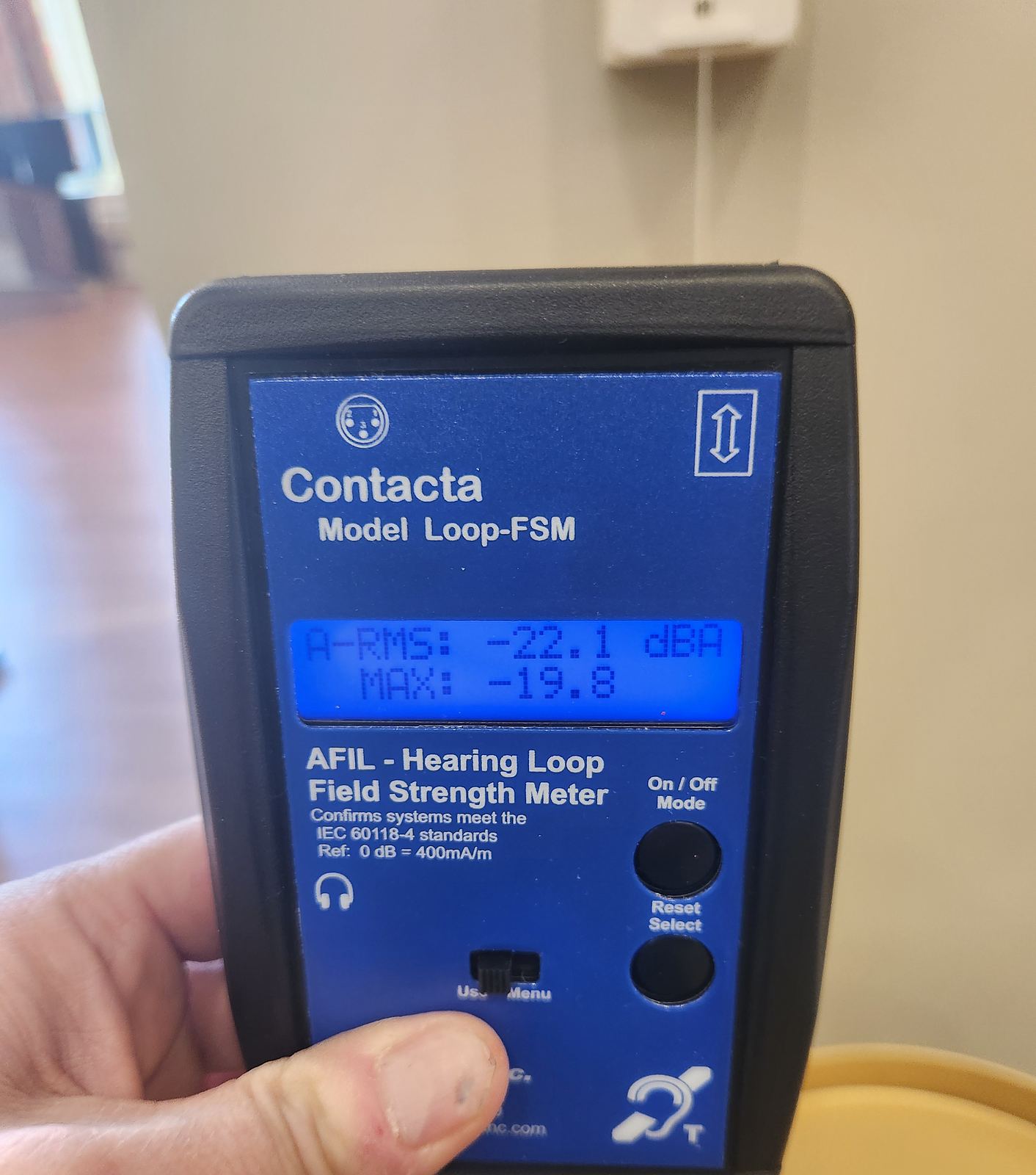 A hand holding a hearing-loop field-strength meter with a live reading on its display