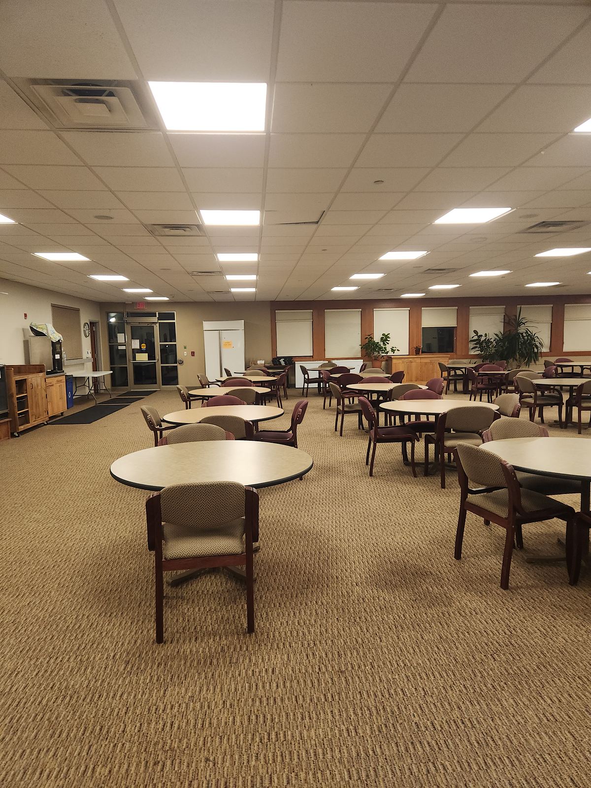 An empty multipurpose room with round tables and upholstered chairs under a lay-in tile ceiling with flat light panels