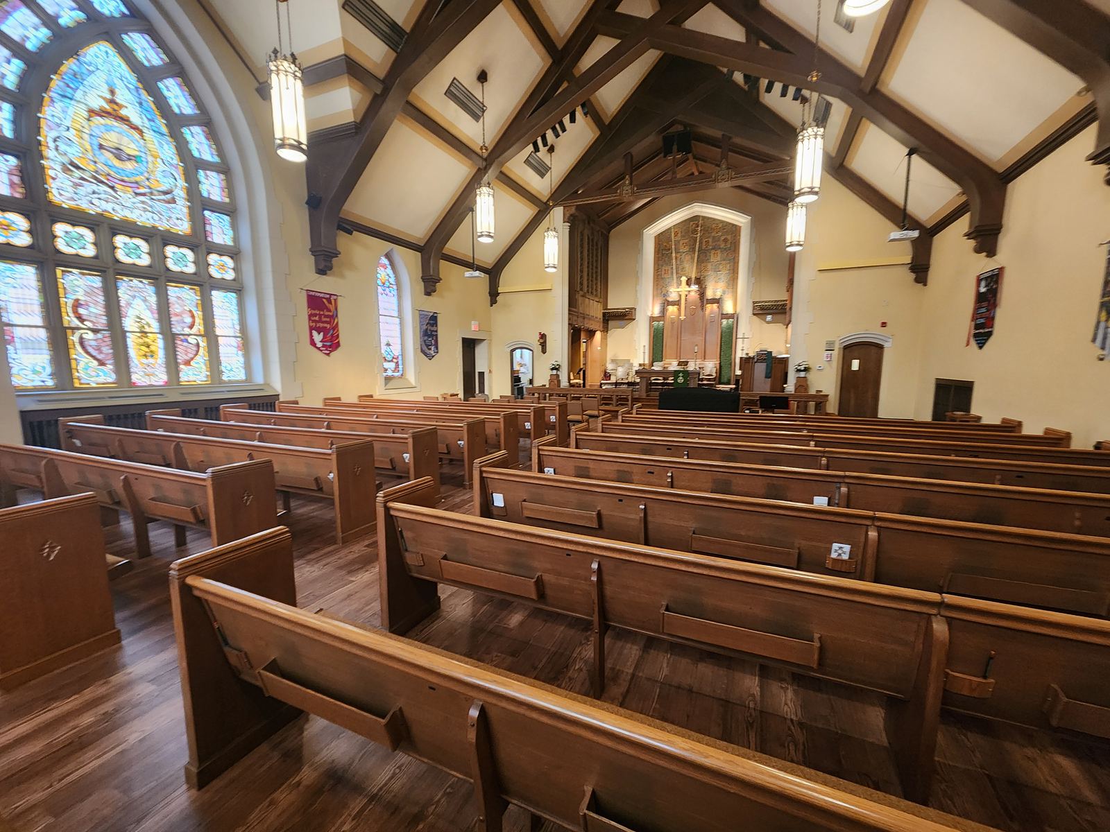 Rows of wooden pews facing the chancel in a sanctuary with tall stained-glass windows and a timber-beamed ceiling